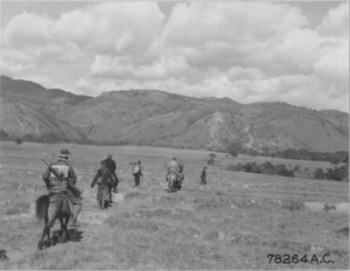 Backs of 16th CCU cameramen riding tiny Chinese horses along path through grassslands on side of small hills.