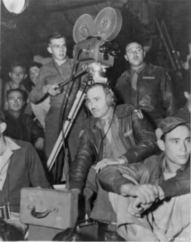 Photo showing several men in U.S. Army uniforms, generally facing the viewer while sitting or standing in an apparently large room and looking toward something beyond the right shoulder of the viewer. Three men are the key figures, as the photo centers on front of a seated man of medium build with slightly receding hairline, in uniform featuring an unzipped, leather bomber jacket with China-Burma-India insignia on the left shoulder. He is wearing 1940s-style headset ear phones with his left hand within an open, medium-sized equipment case, presumably containing sound recording equipment. On our left, behind this man's right shoulder, is a slim young man in wire-frame glasses, standing behind and operating a large motion-picture camera on a tripod stand. Just to the right and slightly behind him, almost directly behind the first man, stands a third man of medium size, wearing an open bomber jacket.  Although it is not apparent that he is doing anything, photo annotation indicates he is the director.  Other men sit on bench seats to either side and behind the three key figures.