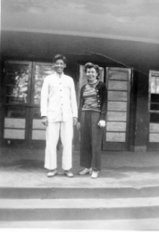 Tall, young Chinese man in white traditional coat and pants stands to the left of Barabara Vatter dressed in loose, dark slacks; darker open, button-front, long sleeve sweater over a horizonatlly striped, light and dark, t-shirt; and light colored, low-heeled shoes.